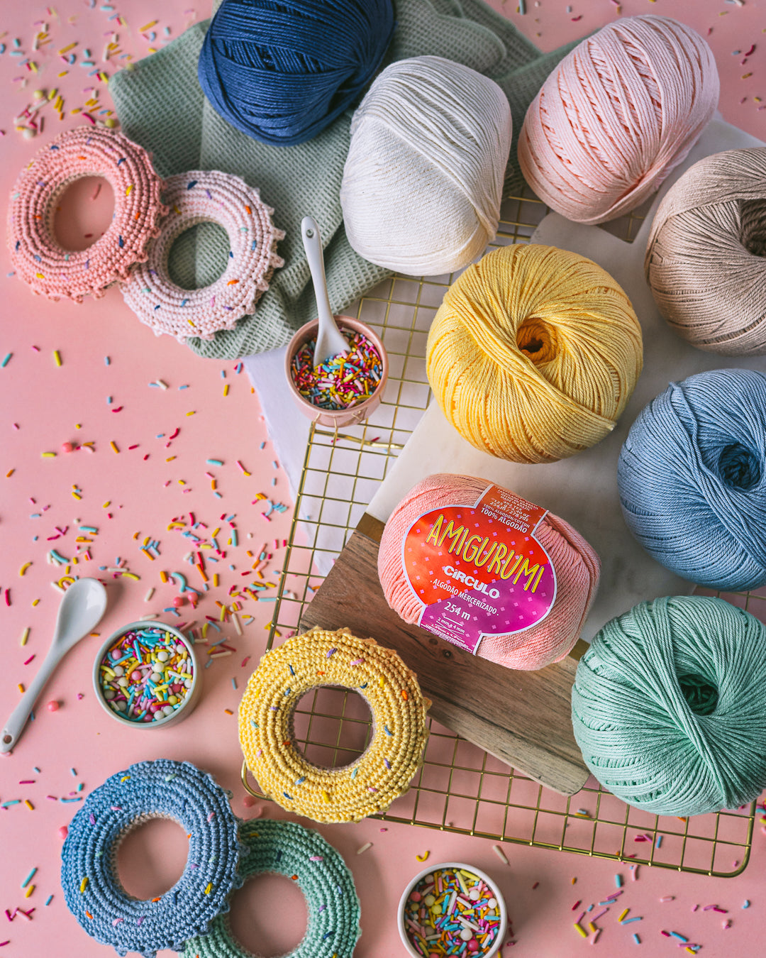 Colorful yarn balls and crocheted donuted on a pink background with sprinkles.