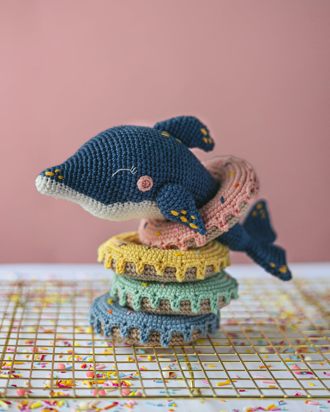 Crocheted dolphin toy on a stack of colorful crocheted donus with a pink background