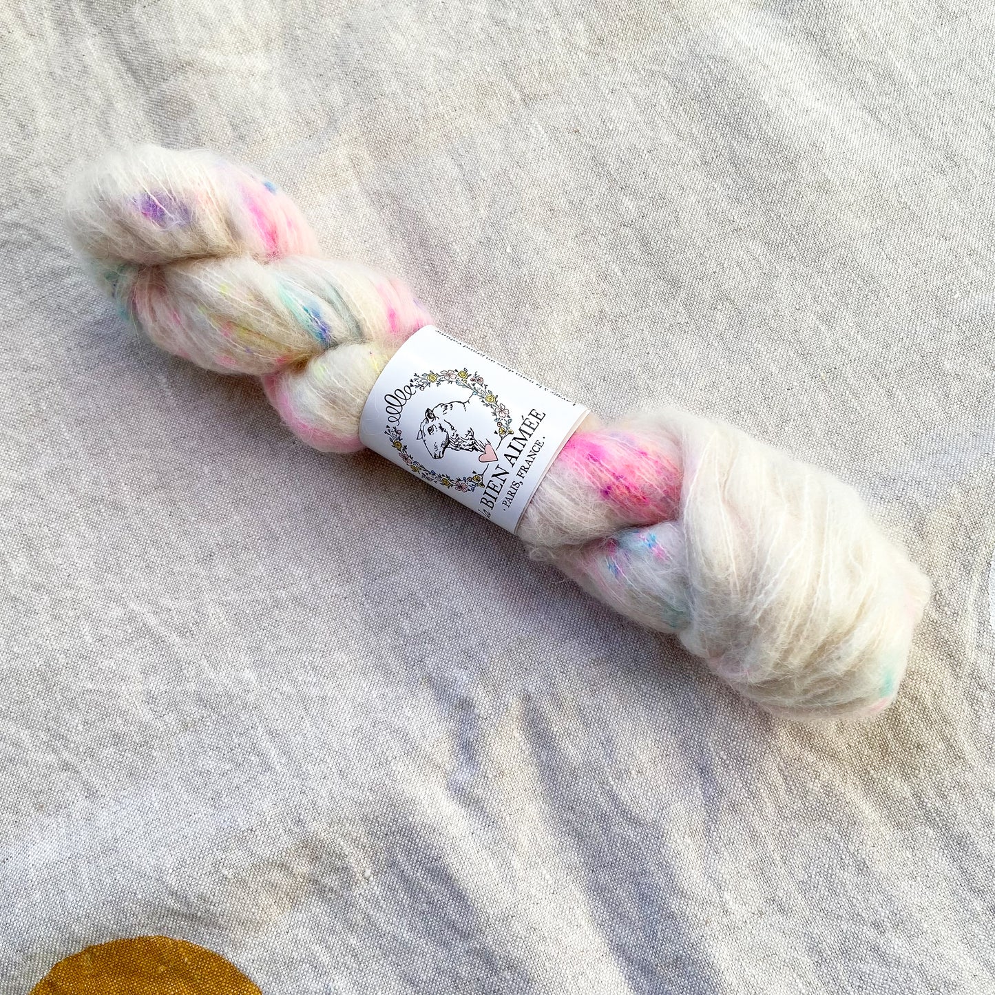 An image of a skein of lace weight yarn in a white base with speckles of pink, yellow, blue, and purple and a white ‘La Bien Aimee’ label against a canvas background.