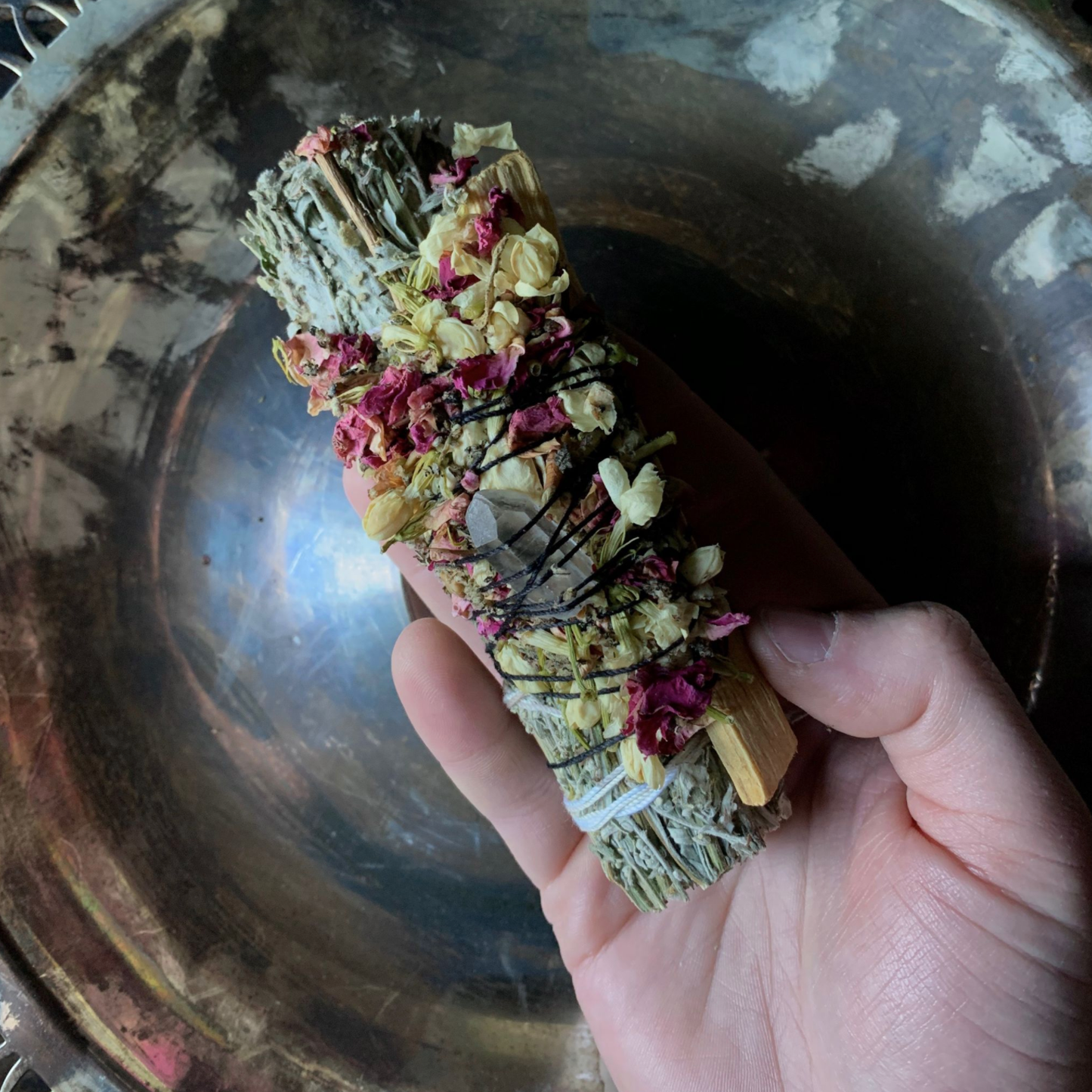 Hand holding a mugwort and palo santo ritual bundle with herbs and flowers over a metallic surface