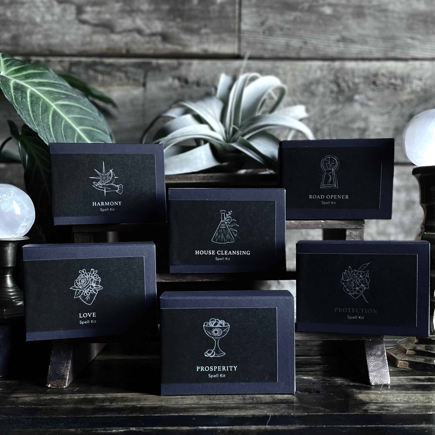 Six spell kits by Ritualcravt arranged on a small wooden shelf: Harmony, House Cleaning, Road Opener, Love, Prosperity and Abundance. The boxes are matte black rectangles with labels. There are plants and crystal balls in the background.