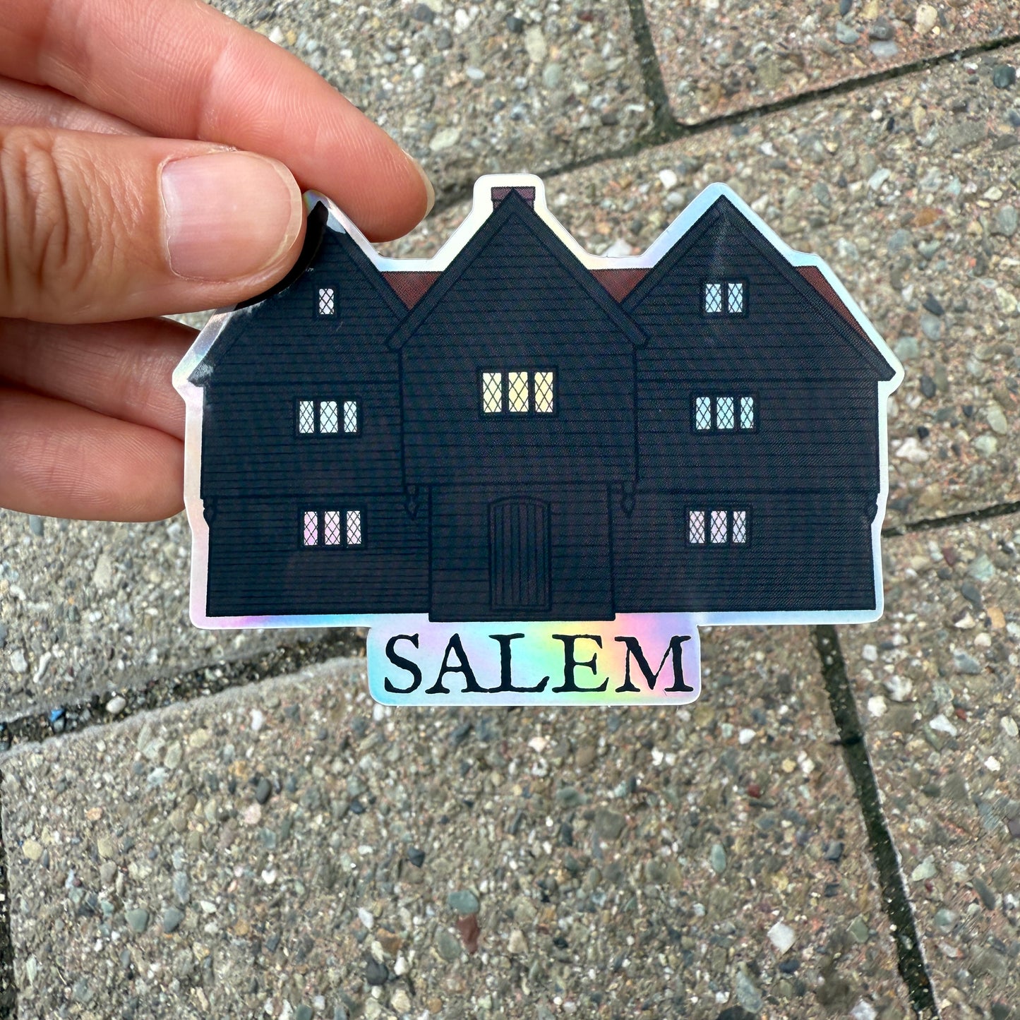 A holographic sticker featuring an illustration of the Witch House in Salem, Massachusetts. A hand is holding the sticker over a stone sidewalk.