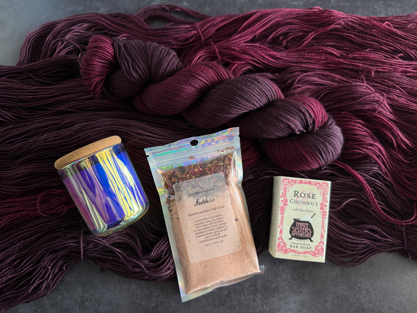 Tonal dark red yarn skein with a shiny glass jar candle, a packet of bath soak, and box of soap laying on yarn on a gray background