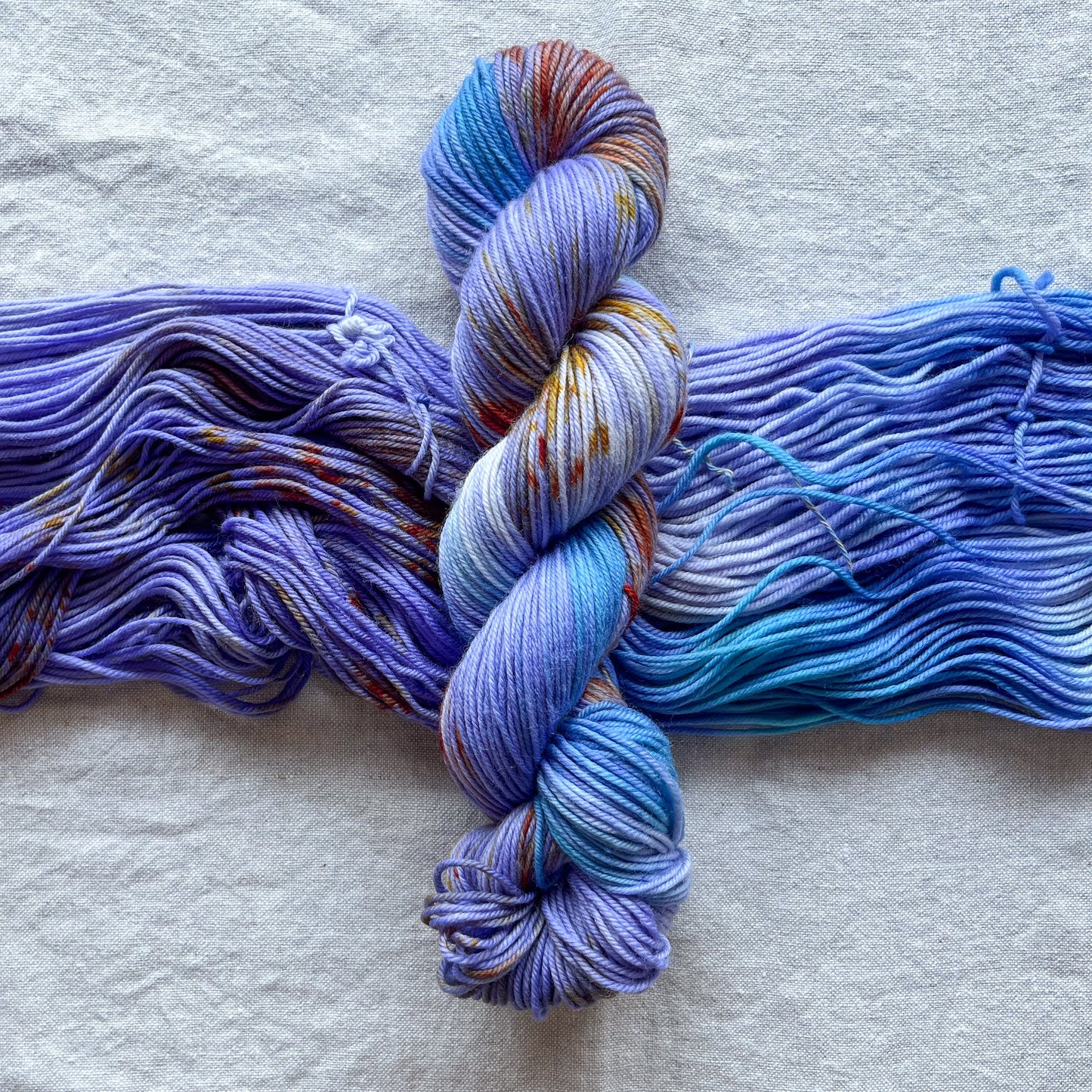 A twisted skein of yarn orientated vertically over an open untwisted skein of yarn. The yarn is a hand painted varigated color way in tones of periwinkle and blue with amber speckles delicately peppered in.