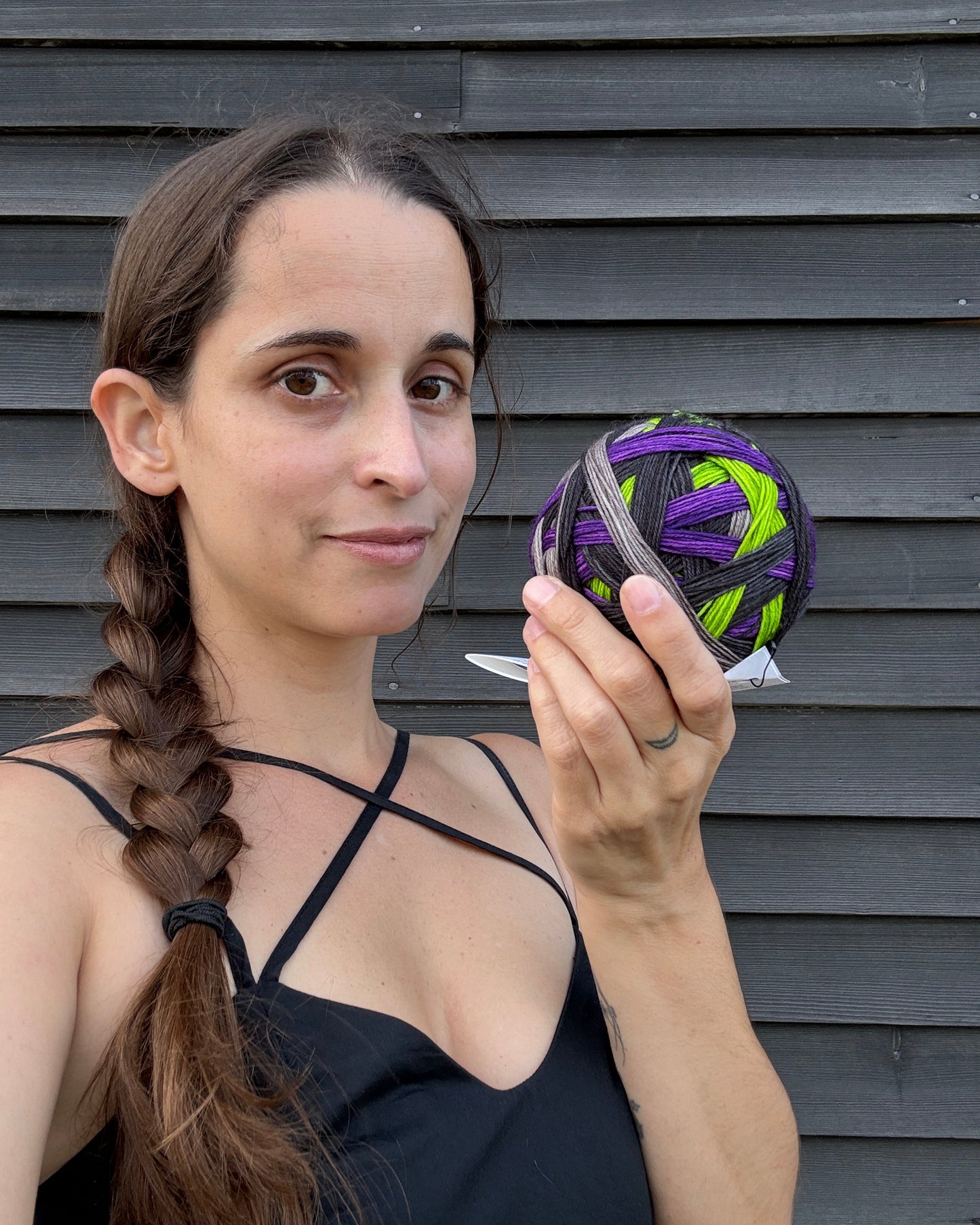 A woman holding up a ball of hand dyed self striping sock yarn in Halloween colors, purple, acid green, black, and grey, against a dark wood background