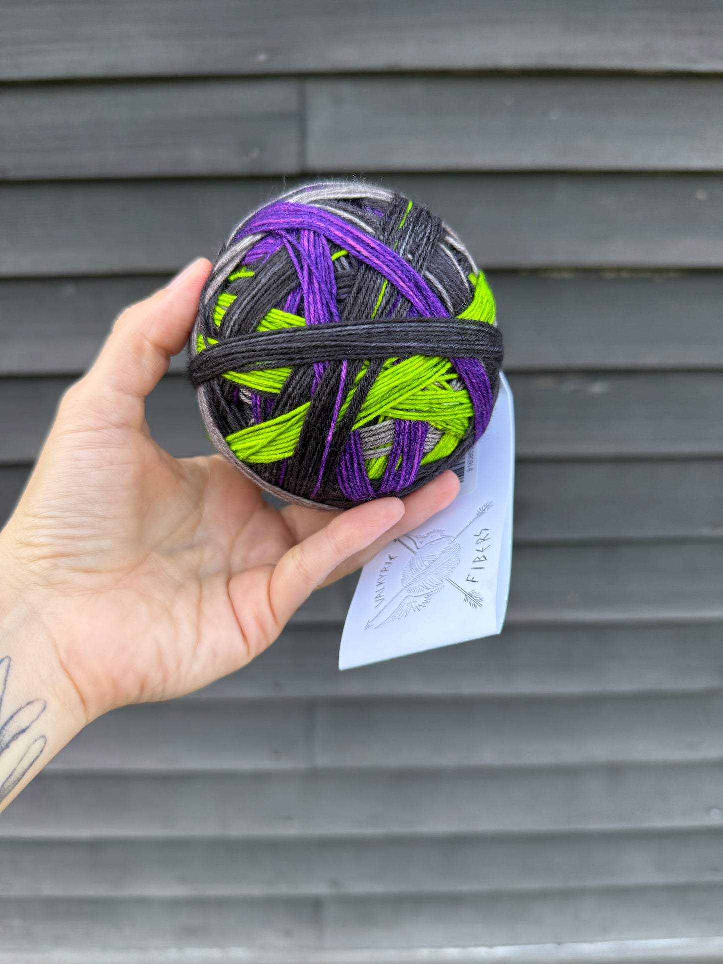 A hand holding up a ball of black, gray, purple, and acid green yarn against a dark wood background