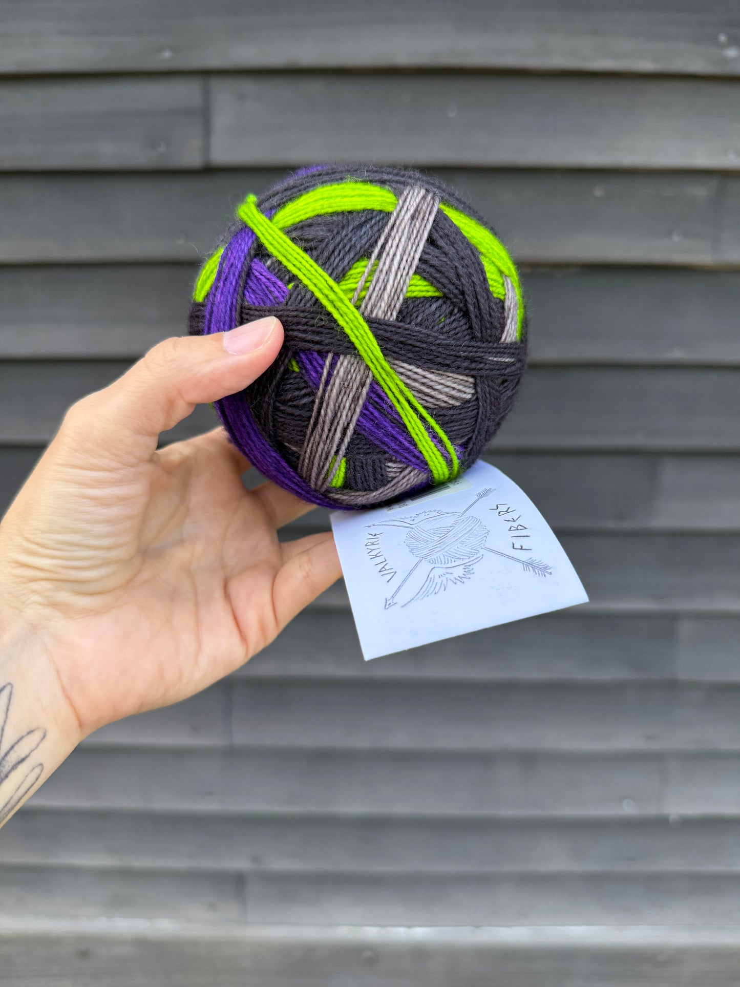 A hand holding up a ball of yarn in acid green, purple, gray and black against a dark wood backgroudn