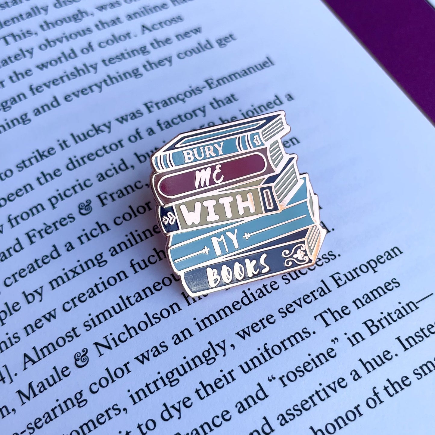 An enamel pin shaped like a stack of books that reads "Bury Me With My Books" on a page of a book.