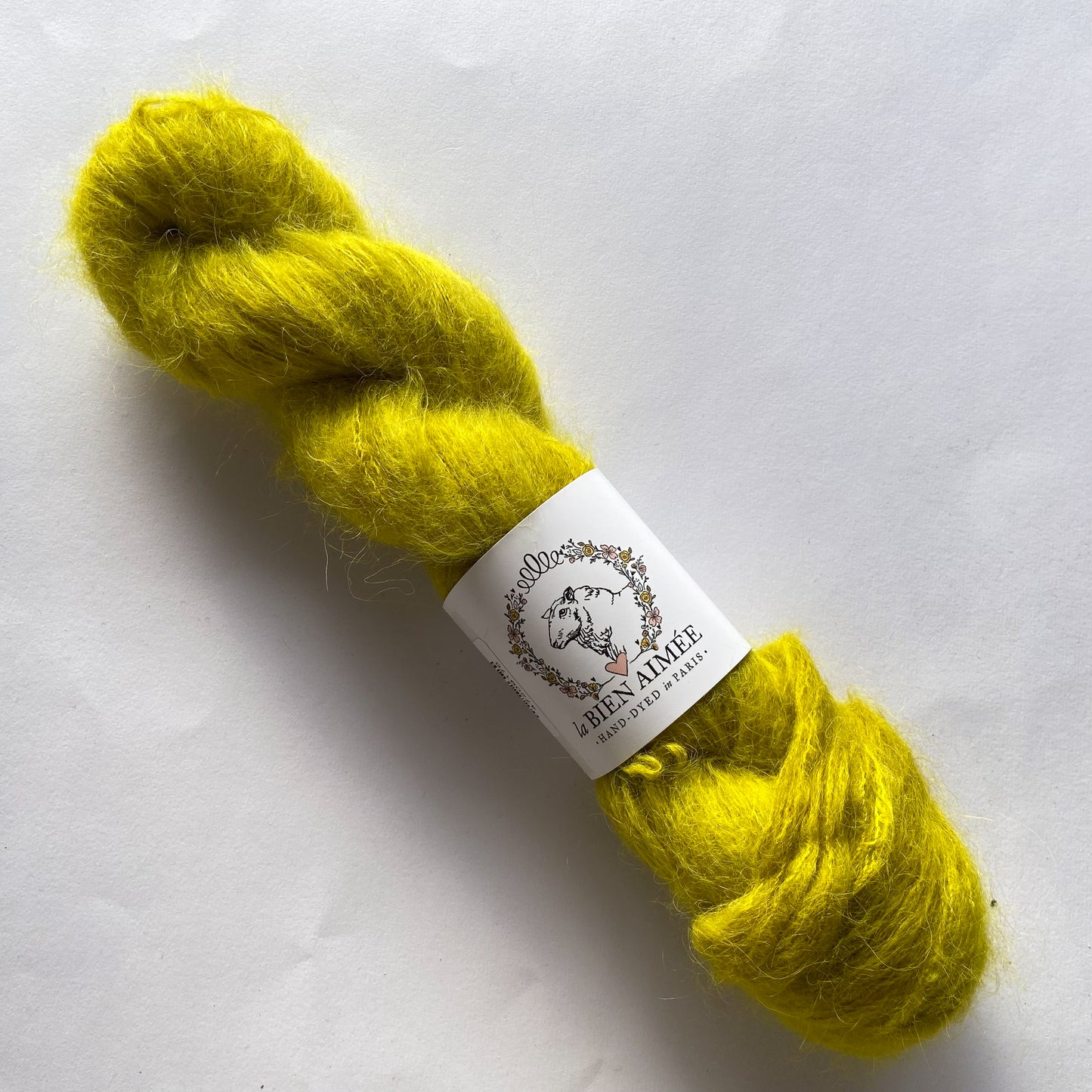 An image of a skein of La Bien Aimee lace weight yarn in a bright yellow green color with a white ‘La Bien Aimee’ label against a white background.