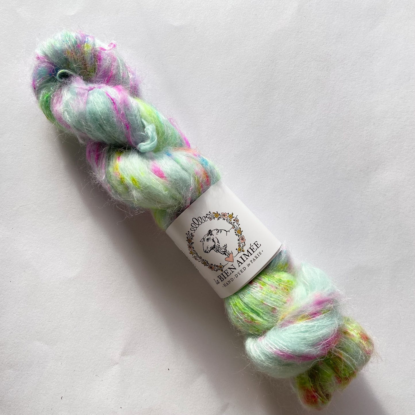 An image of a skein of lace weight yarn in a pale blue base with speckles of neon green and pink with a white ‘La Bien Aimee’ label against a white background.