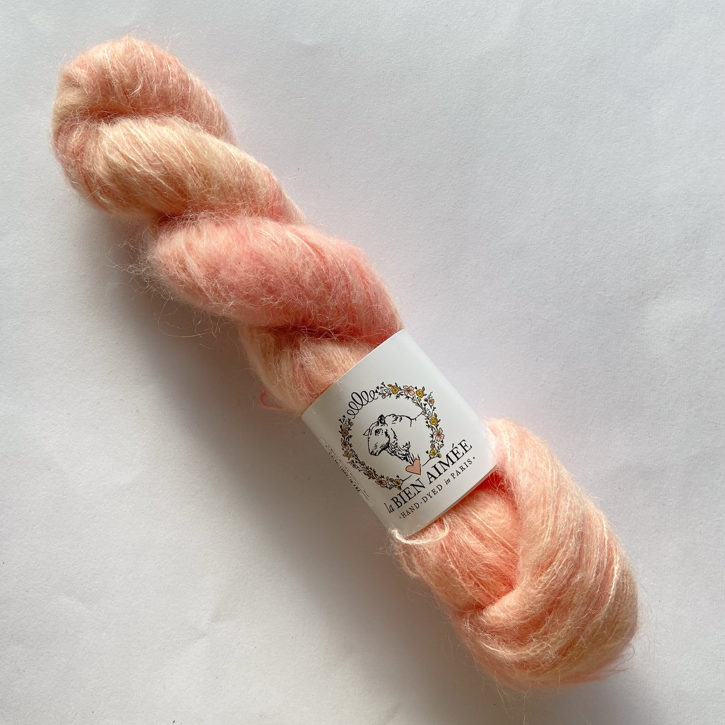 An image of a skein of lace weight yarn in shades of peach pink with a white ‘La Bien Aimee’ label against a white background.