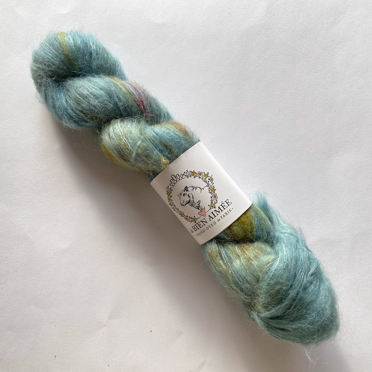 An image of a skein of lace weight yarn in light blue, yellow, and pink colors with a white ‘La Bien Aimee’ label against a white background.