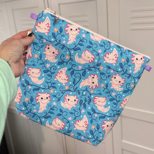 Zippered knitting project with a cute axolotl pattern ob a blue background held by a hand against a white background