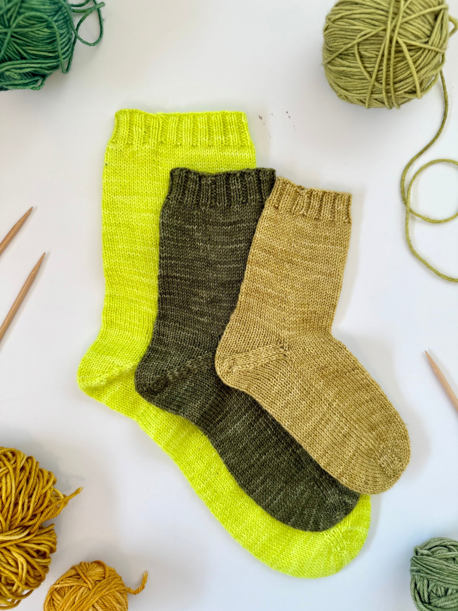 Three knitted socks in green, brown, and beige with yarn and knitting needles on a white background.