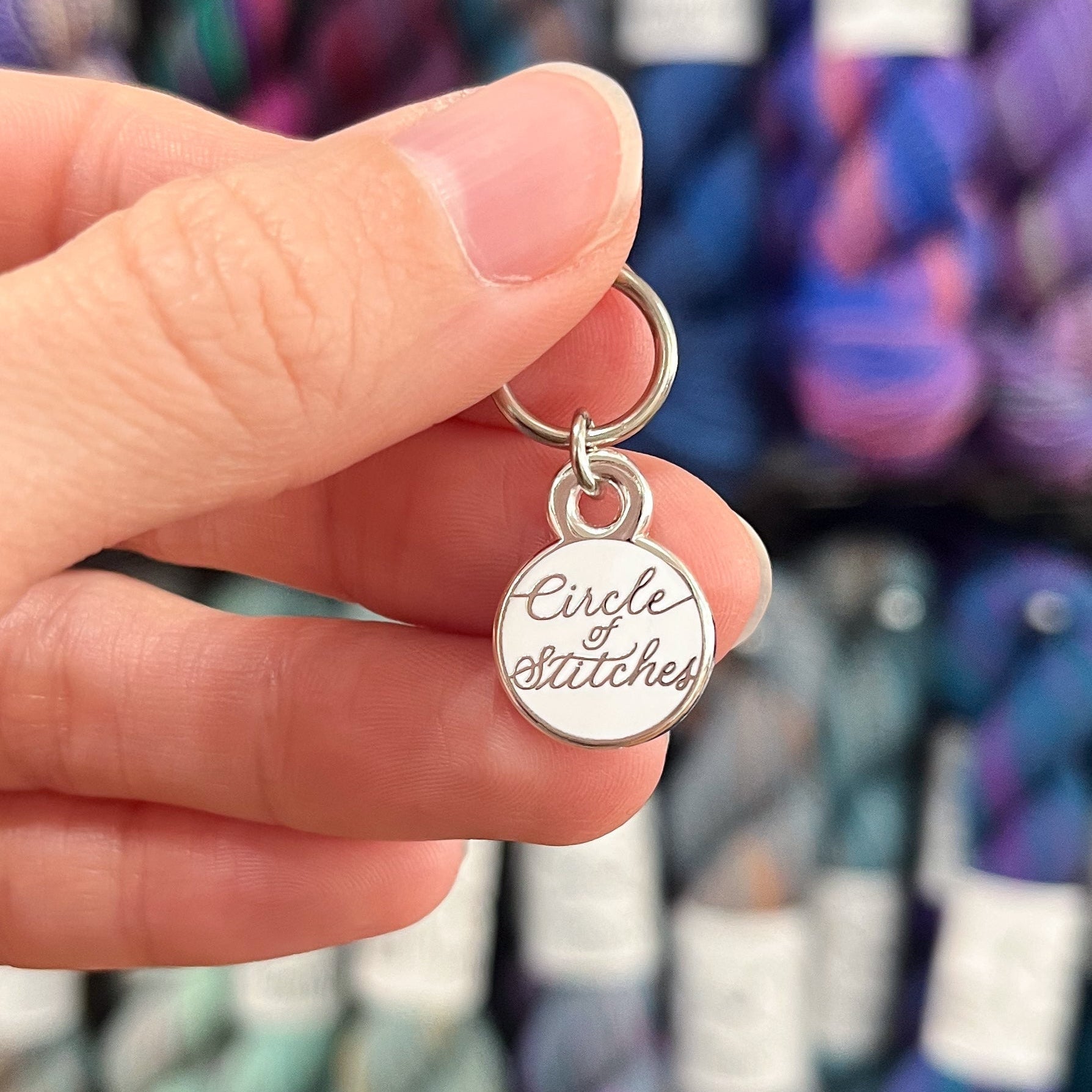 Hand holding a silver charm with 'Circle of Stitches' text against a blurred yarn background