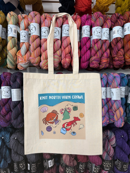 A tote bag featuring a lobster, crab, and shrimp knitting and crocheting design on a beach,  with the text 'Knit North Yarn Crawl on a background of colorful yarn skeins.
