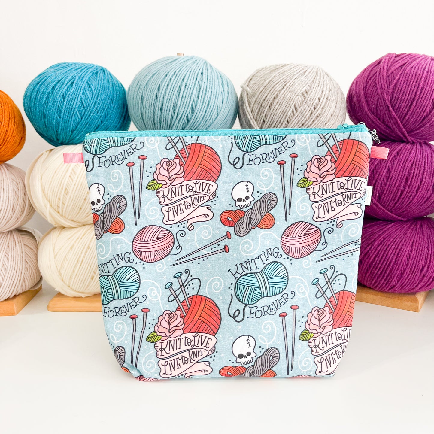 A light blue patterned project bag with knitting-themed elements and skulls, placed among various colored yarns.