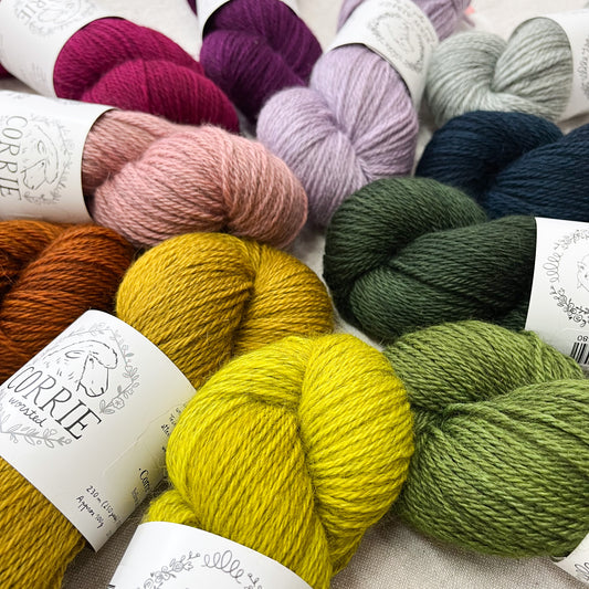 An image of multiple skeins of La Bien Aimee worsted weight yarn in a variety of colors with white labels featuring an image of a sheep and the title ‘Corrie Worsted’ against a canvas background.