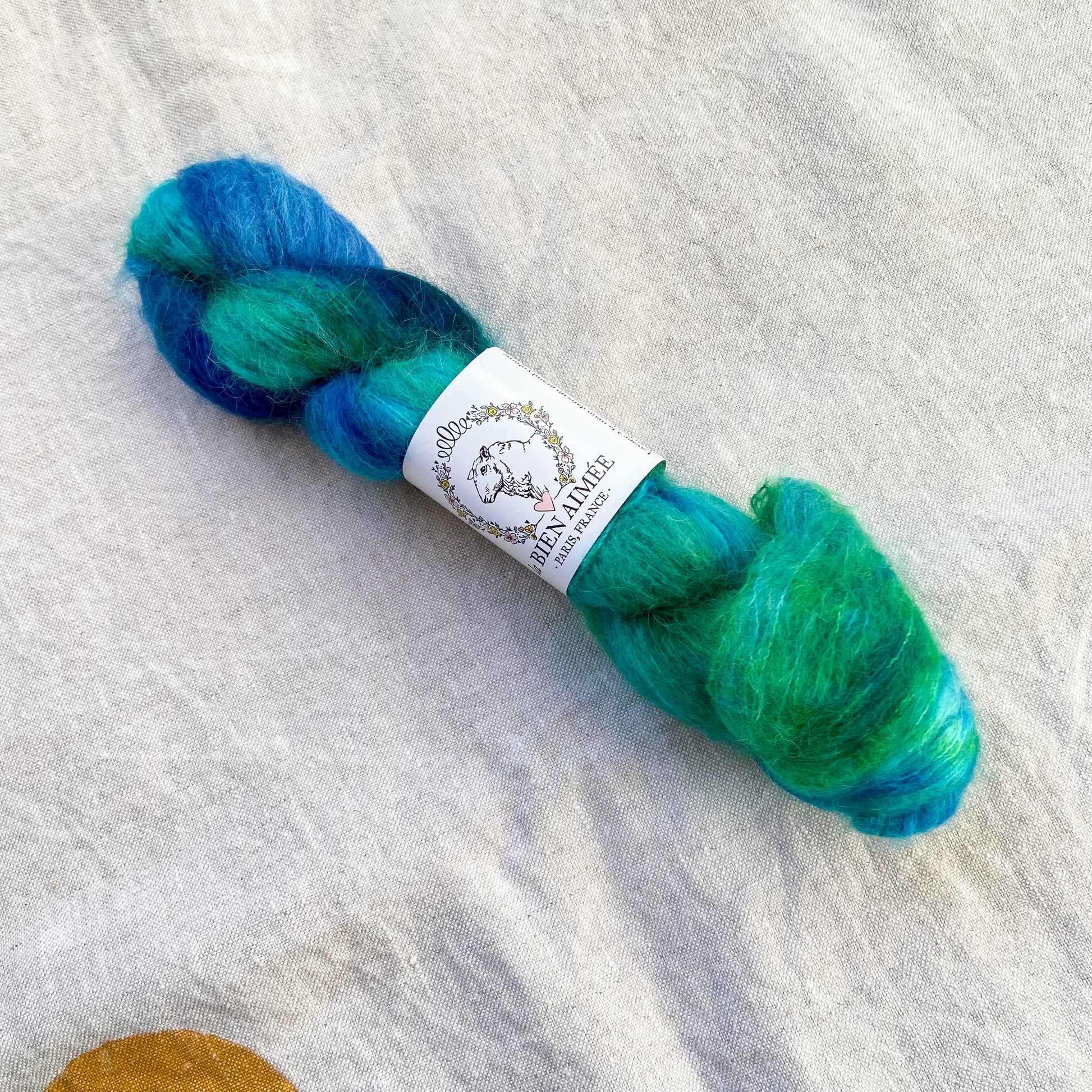 An image of a skein of lace weight yarn in vibrant shades of blue and green with a white ‘La Bien Aimee’ label against a canvas background.