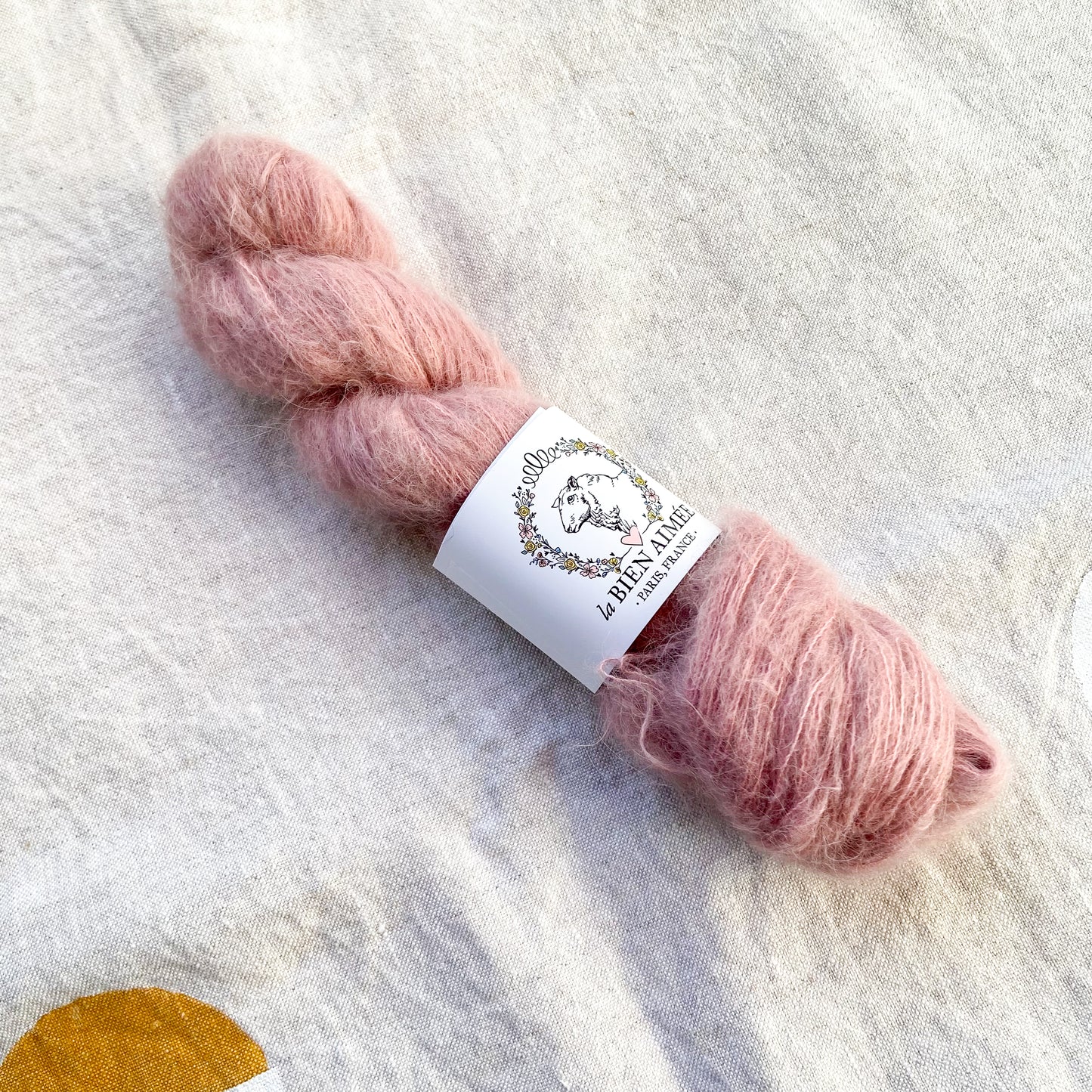 An image of a skein of lace weight yarn in a dusty pink color with a white ‘La Bien Aimee’ label against a canvas background.