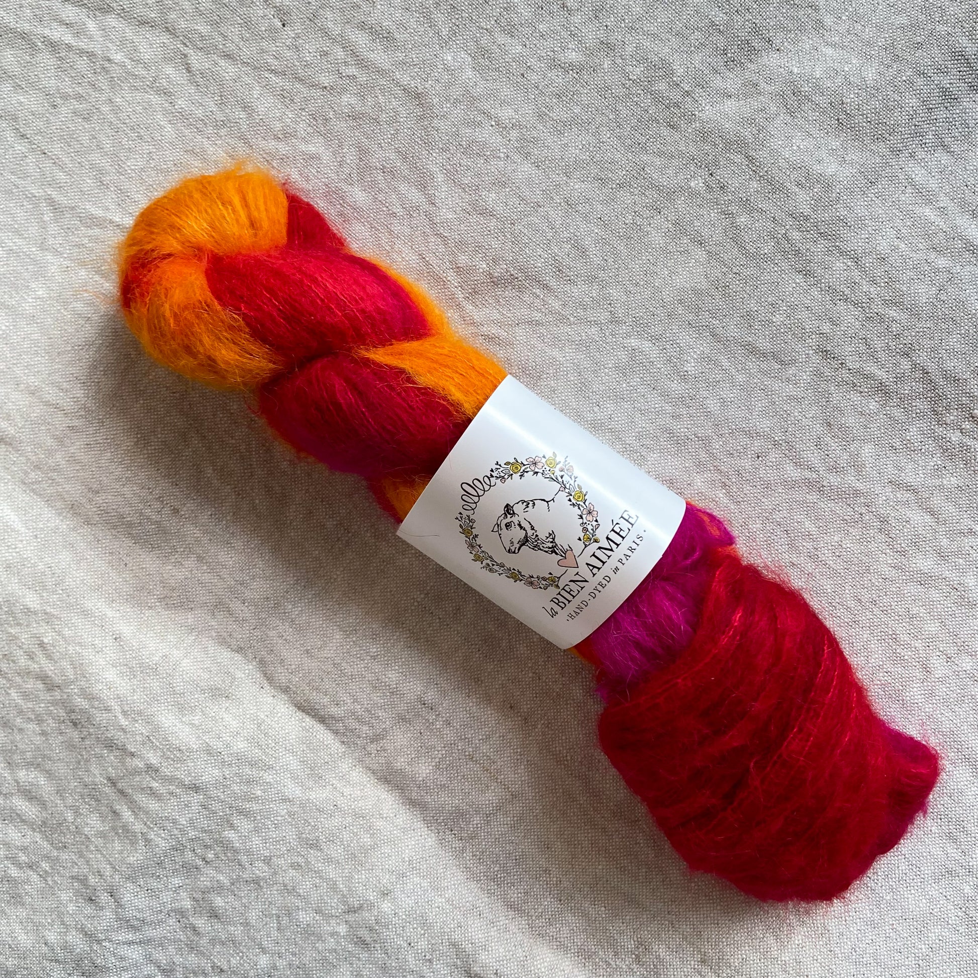 An image of a skein of lace weight yarn in vibrant red, orange and pink colors with a white ‘La Bien Aimee’ label against a canvas background.