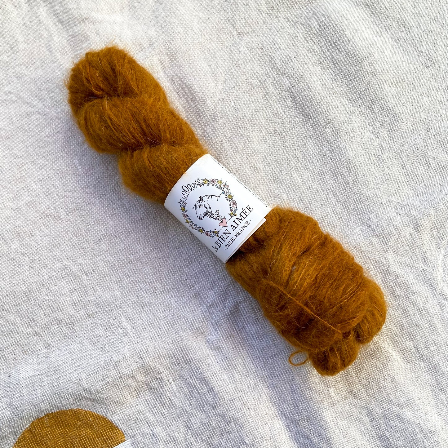 An image of a skein of lace weight yarn in a light brown color with a white ‘La Bien Aimee’ label against a canvas background.