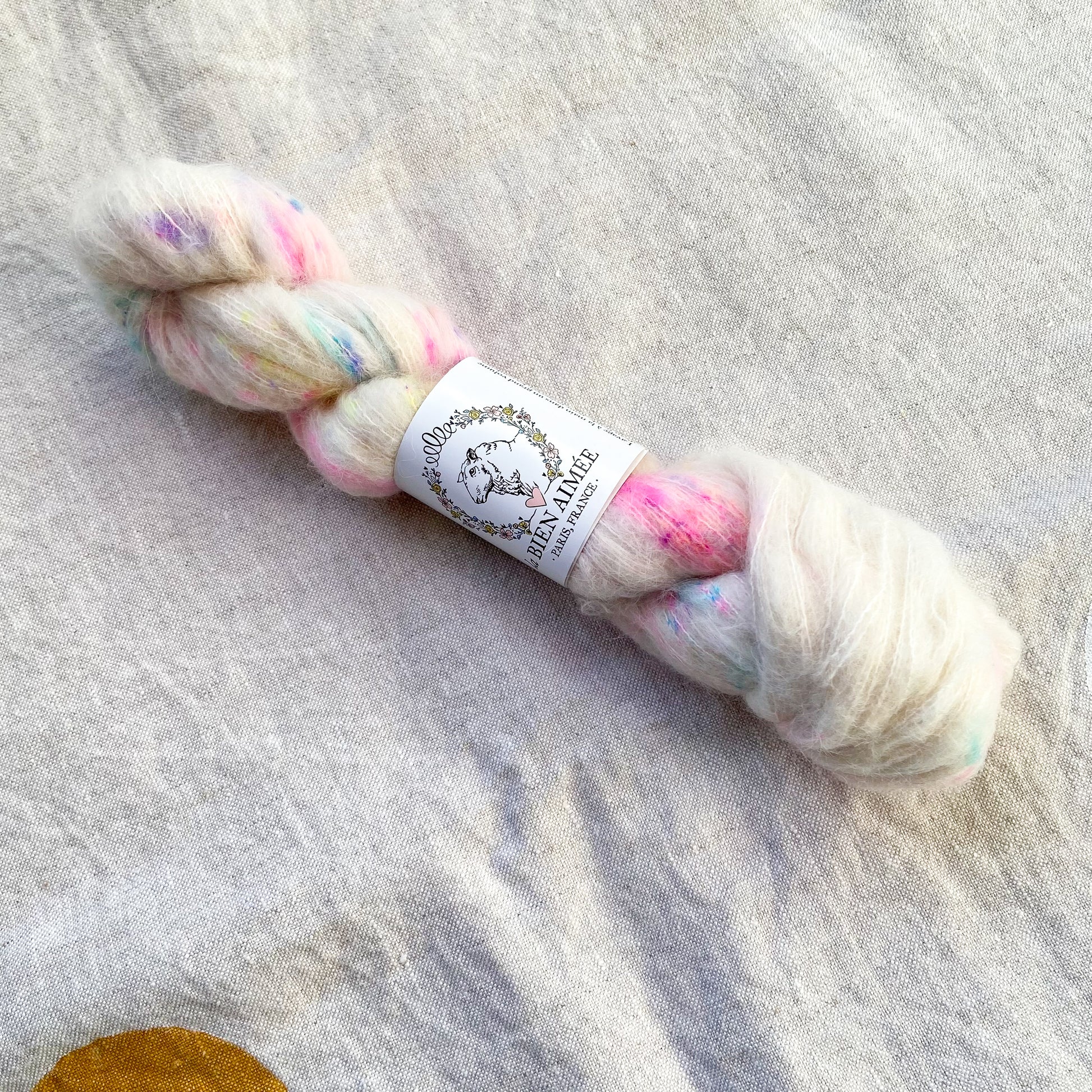 An image of a skein of lace weight yarn in a white base with speckles of pink, yellow, blue, and purple and a white ‘La Bien Aimee’ label against a canvas background.