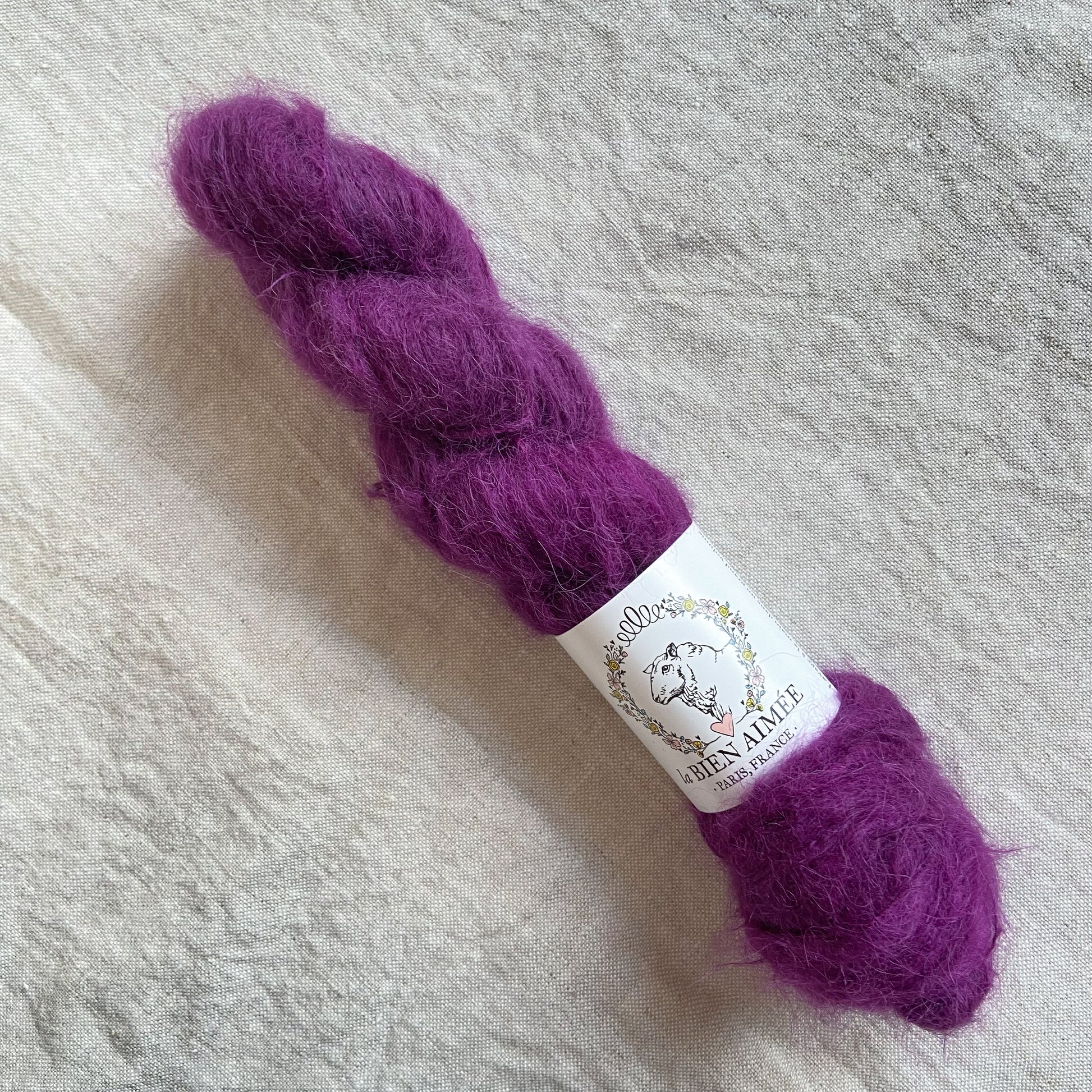 An image of a skein of lace weight yarn in the color purple with a white ‘La Bien Aimee’ label against a canvas background.