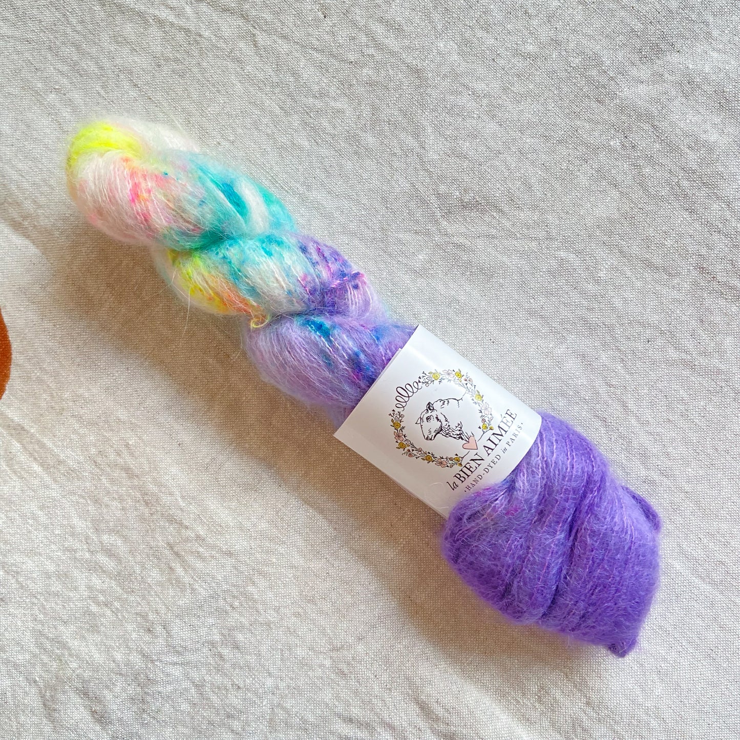 An image of a skein of lace weight yarn in purple and white with  blue and yellow speckles and a white ‘La Bien Aimee’ label against a canvas background.