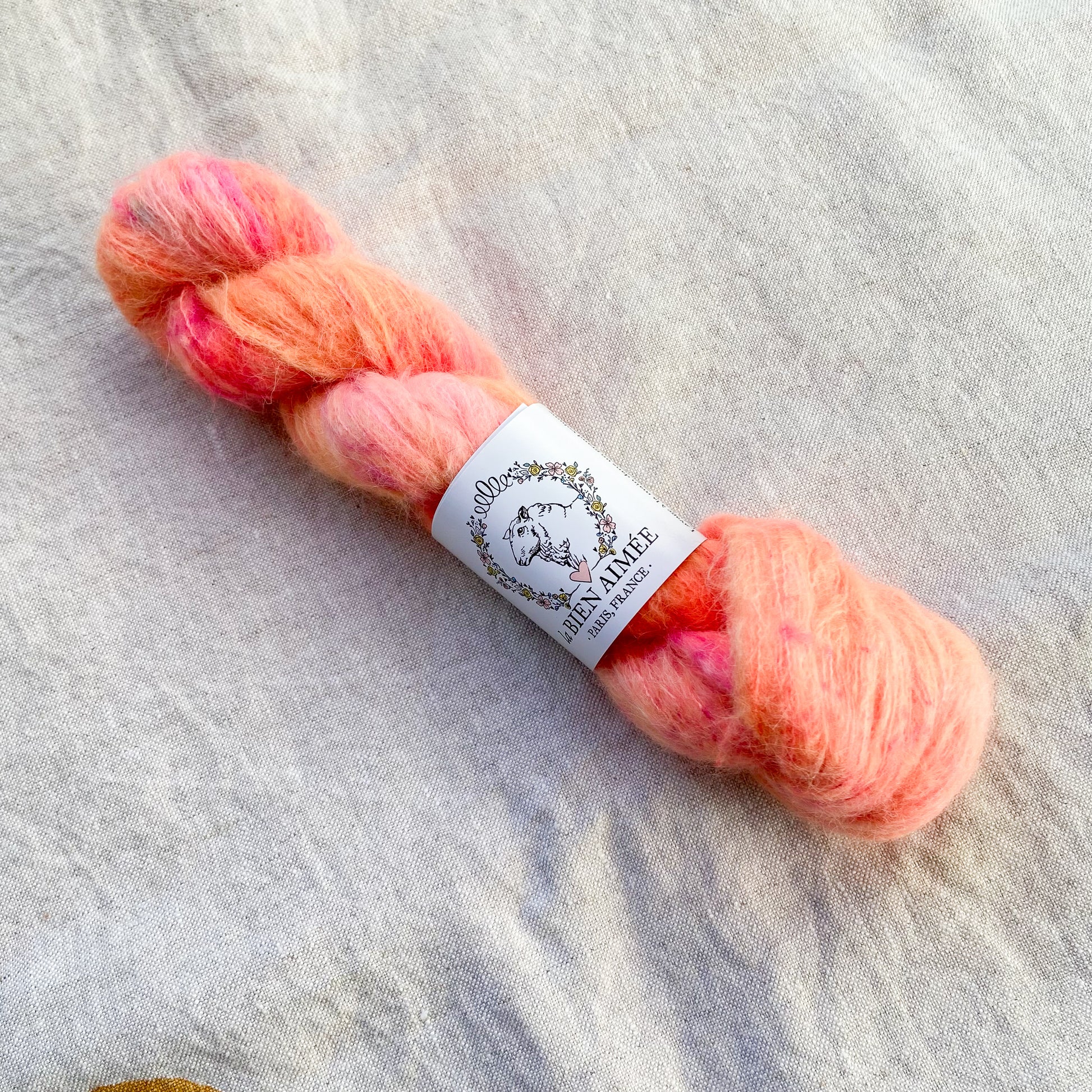 An image of a skein of lace weight yarn in vibrant shades of peach pink and orange with a white ‘La Bien Aimee’ label against a canvas background.