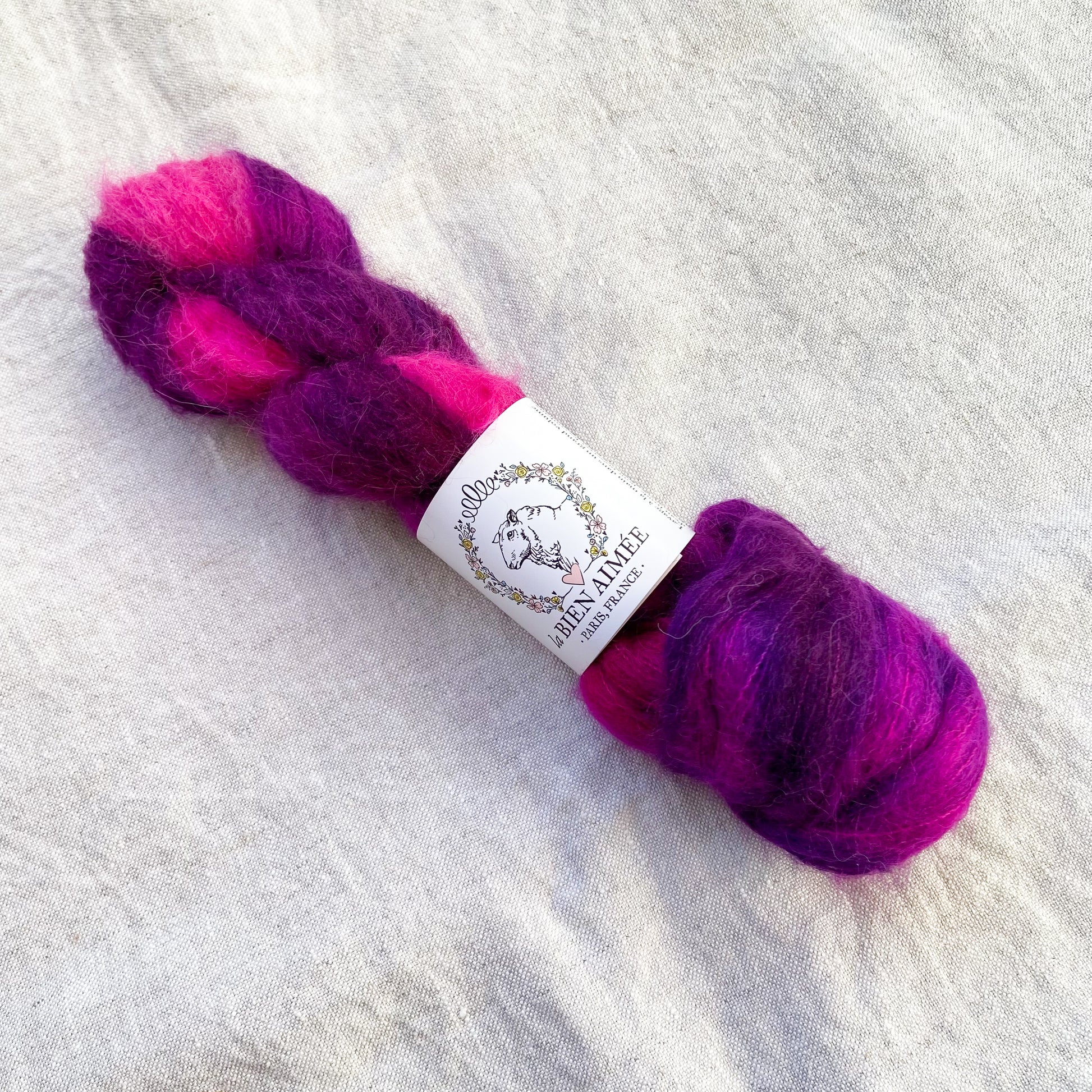 An image of a skein of lace weight yarn in vibrant shades of pink and purple with a white ‘La Bien Aimee’ label against a canvas background.