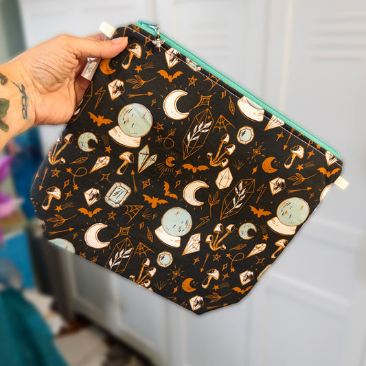 A hand holding a wedge bag made with black fabric with a witchy print featuring crystal balls, crescent moons, bats, mushrooms, crystals, and spider webs, with an aqua zipper