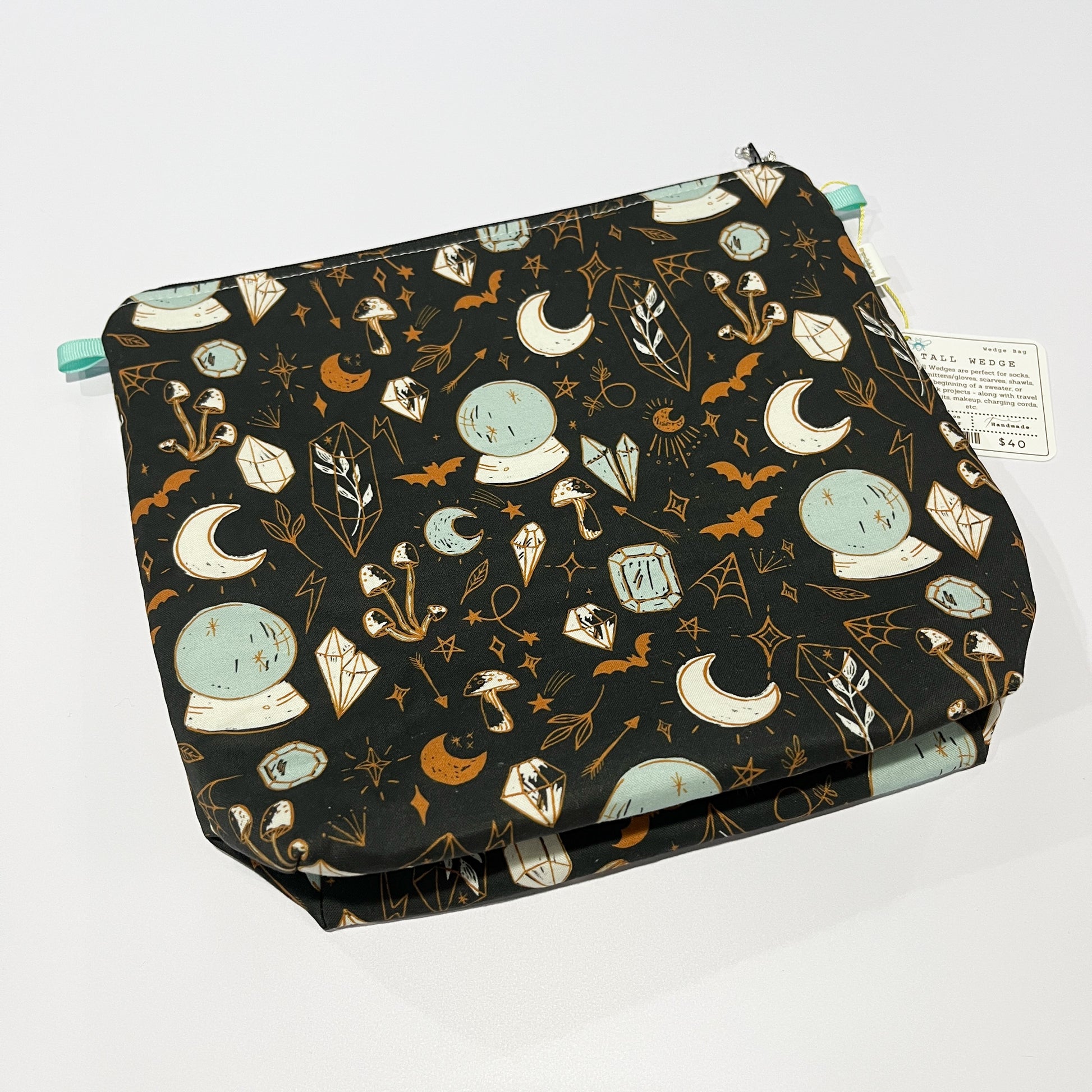 A tall wedge-shaped project bag with a multicolored pattern including crystal balls, moons, and other whimsical motifs, closed with an aqua zipper.