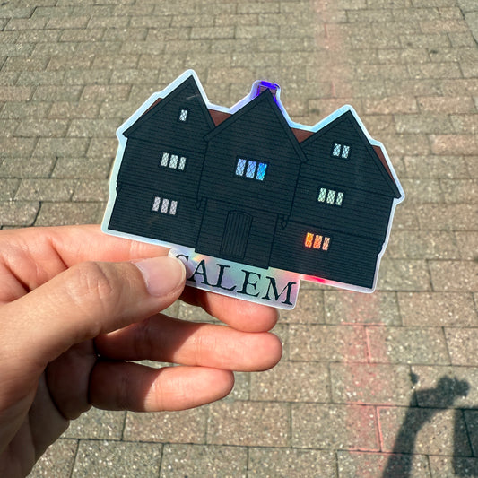 A hand holding up a sticker over a stone sidewalk, the sticker is an illustration of The Witch House in Salem, Massachusetts. It is a house with three gables and black siding. Underneath the house is the word "Salem" The sticker is printed on holographic vinyl. 