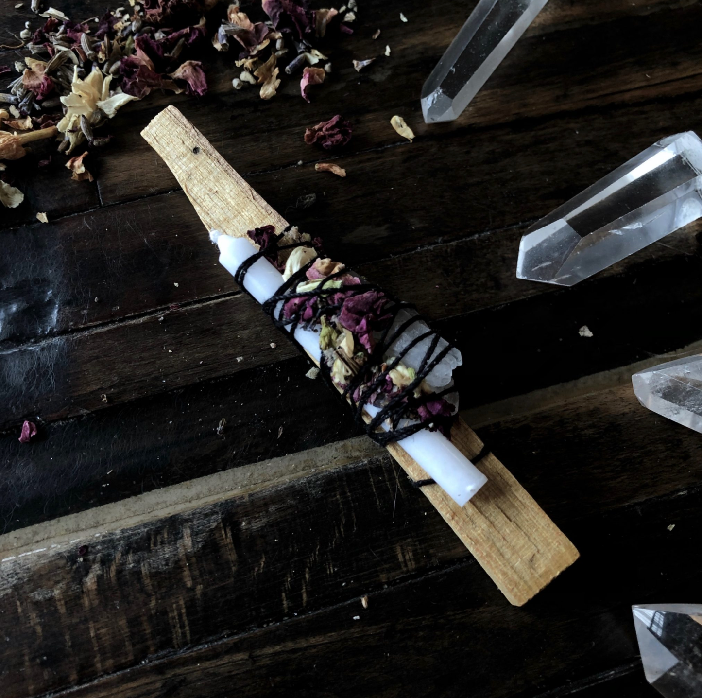 A spell bundle for intention setting by Ritualcravt, with palo santo, a white, candle, crystal, and herbs, tied together with black string, on a wooden table surrounded by crystals and herbs