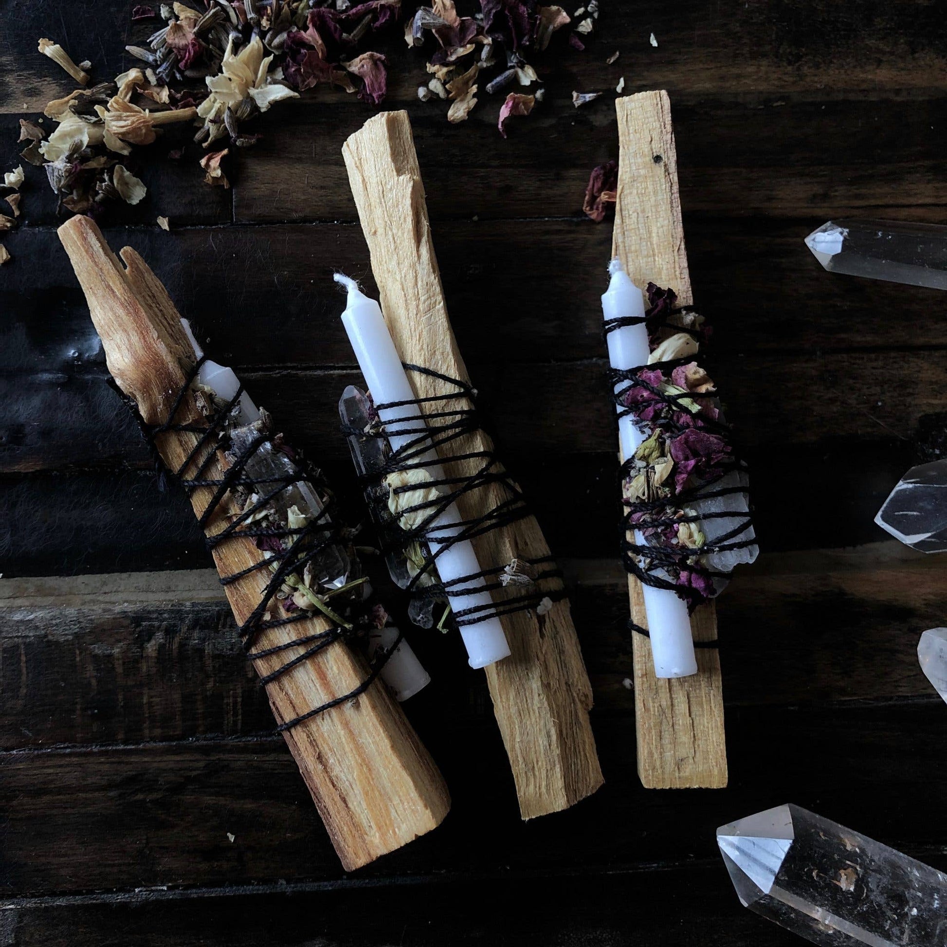 Three spell bundles with Palo Santo sticks, herbs, and crystals arranged on a wooden surface, with white candles and black string bindings.