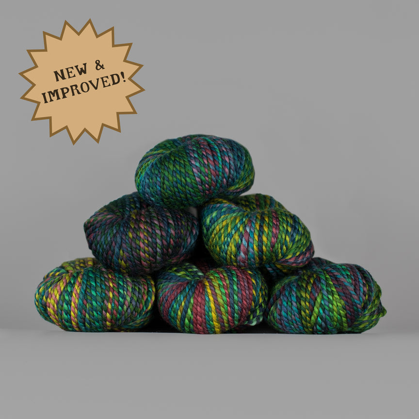 Five skeins of variegated yarn stacked in a pyramid on a gray background, with a tan starburst badge in the top-left corner reading 'NEW & IMPROVED!'. The yarn displays a multicolor variegated pattern with shades of green, blue, purple, yellow, and pink.