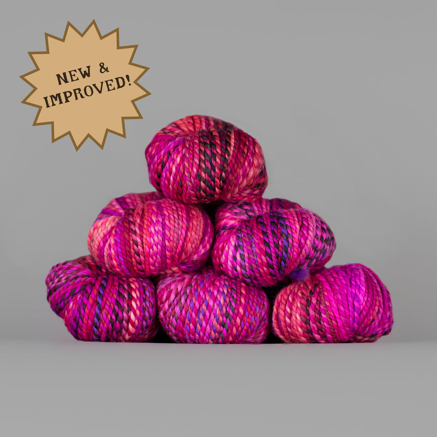 A stack of six variegated yarn balls in vibrant shades of magenta, pink, and purple with dark accents, set against a solid gray background. A gold starburst badge in the top-left corner reads 'NEW & IMPROVED!'.