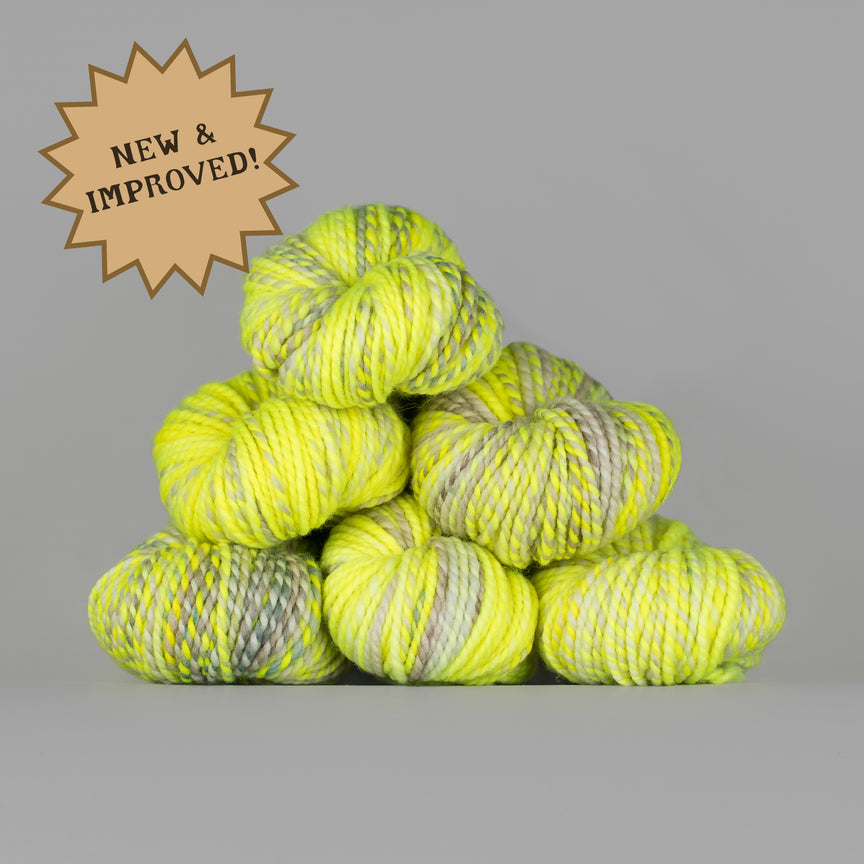 A stack of bright neon yellow and gray variegated yarn skeins on a solid gray background, with a brown starburst badge in the top-left corner reading 'NEW & IMPROVED!'.