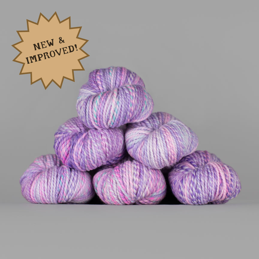 A stack of eight variegated yarn skeins in shades of purple, pink, and blue, set against a solid gray background with a gold starburst badge in the top left corner reading 'NEW & IMPROVED!'.