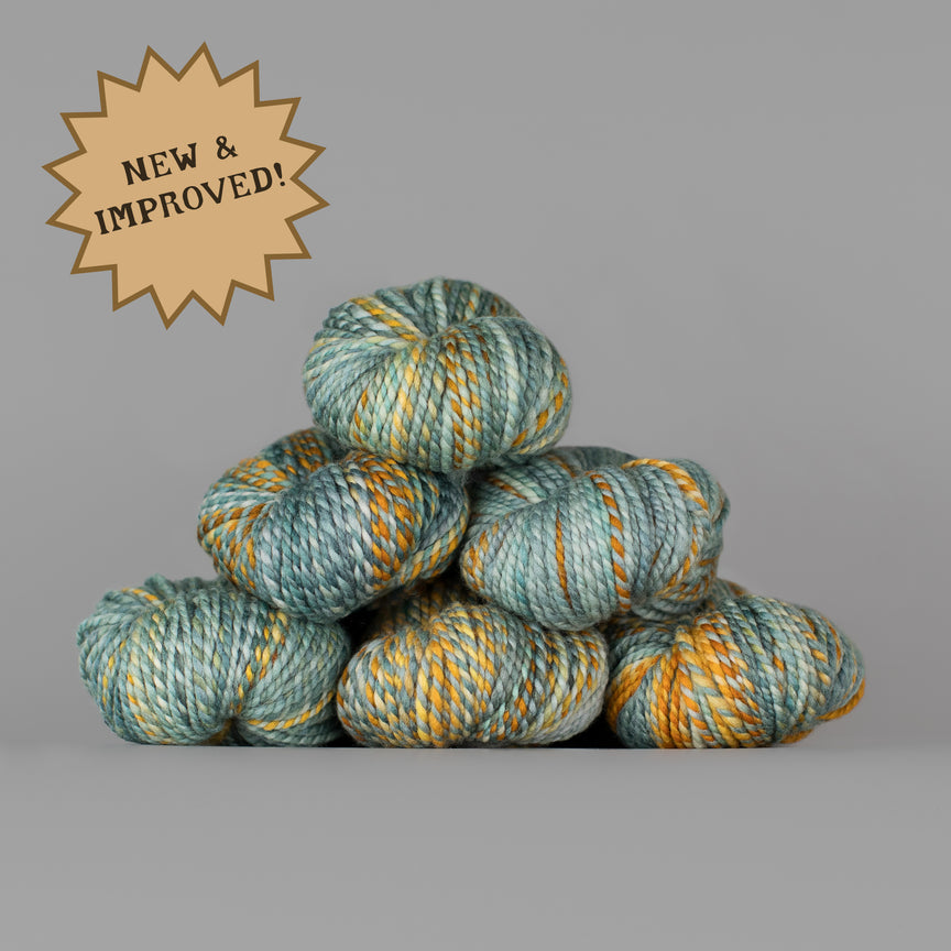 A stack of variegated yarn skeins in muted teal, sage green, and mustard yellow on a solid gray background, with a tan starburst badge in the top-left corner reading 'NEW & IMPROVED!'.