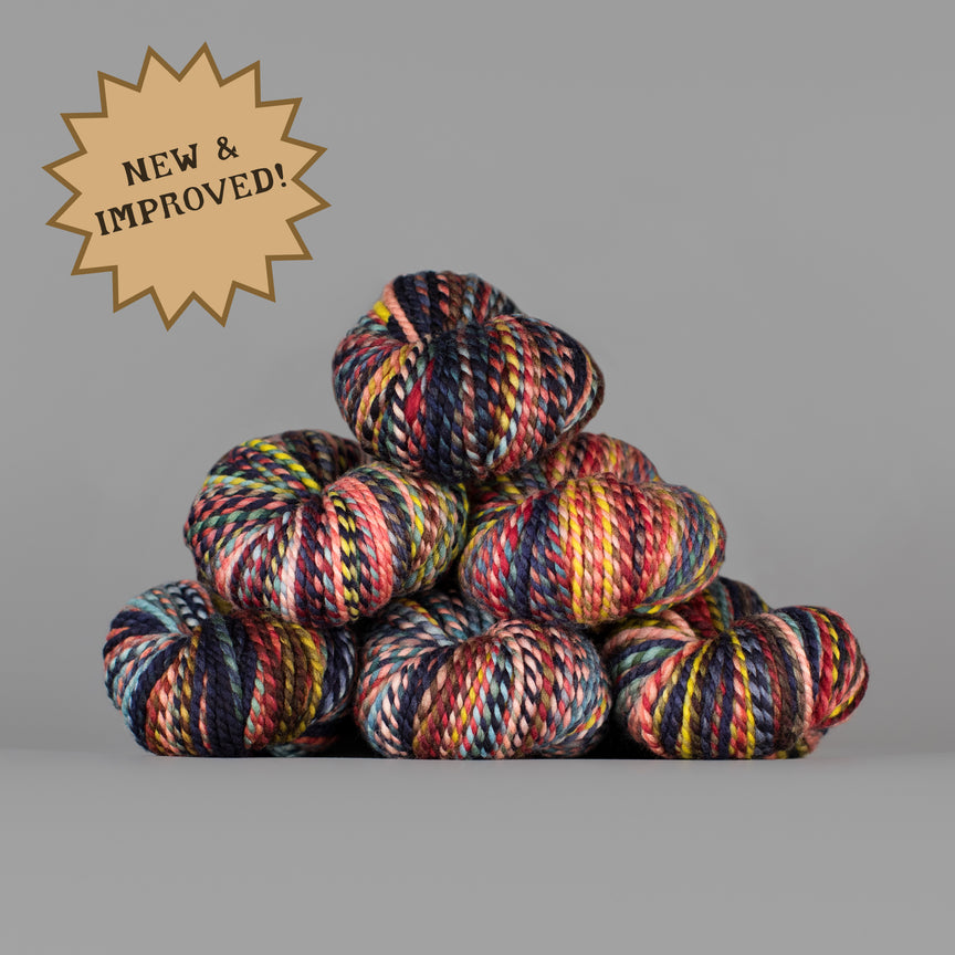 A stack of multicolored variegated yarn skeins on a plain gray background, with a tan starburst badge in the upper left reading 'NEW & IMPROVED!'.