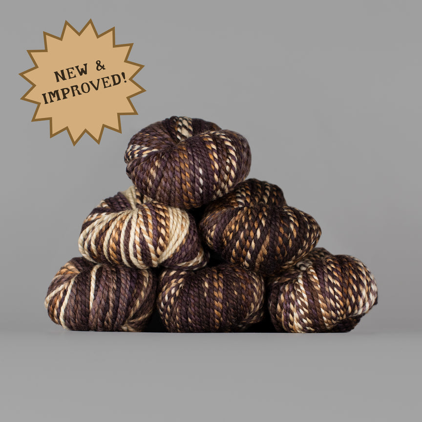 A stack of nine variegated yarn skeins in dark brown, black, and cream, with a tan starburst badge in the top-left corner reading 'NEW & IMPROVED!'. The background is a solid neutral gray.