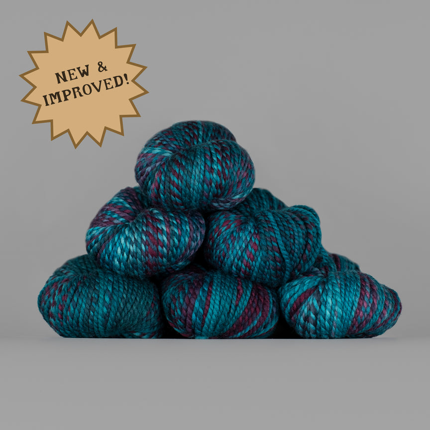 A stack of variegated yarn skeins in teal, blue, and purple tones on a plain gray background, with a tan starburst badge in the upper left reading 'NEW & IMPROVED!'.