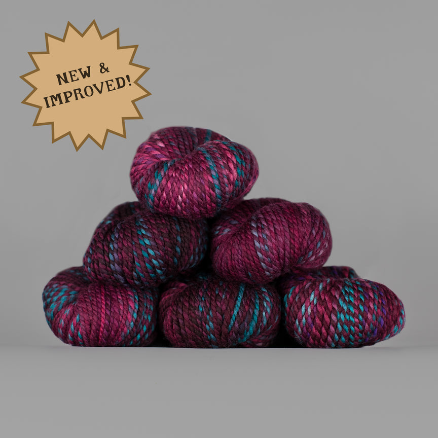 A stack of six variegated yarn skeins in shades of magenta, purple, and teal on a solid gray background, with a gold starburst badge in the top-left corner reading 'NEW & IMPROVED!'.