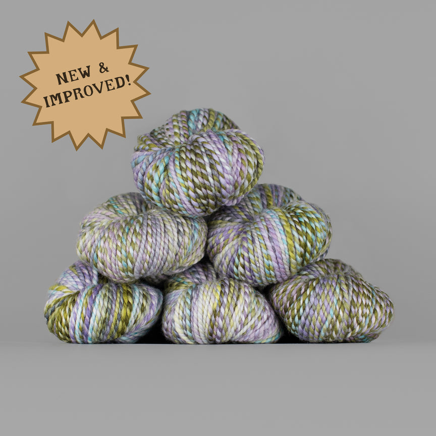 A stack of variegated yarn skeins in pastel green, lavender, and cream tones on a solid gray background, with a tan starburst badge in the top-left corner reading 'NEW & IMPROVED!'.
