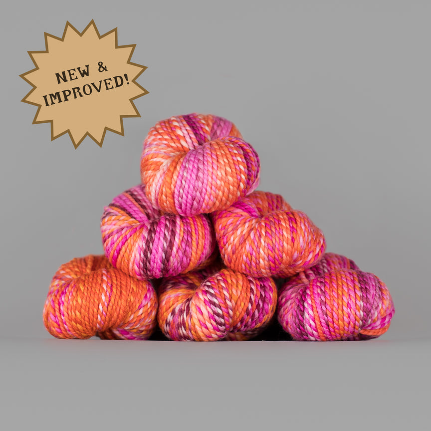A stack of variegated yarn skeins in bright orange, pink, and purple tones on a plain gray background, with a tan starburst badge in the upper left reading 'NEW & IMPROVED!'.