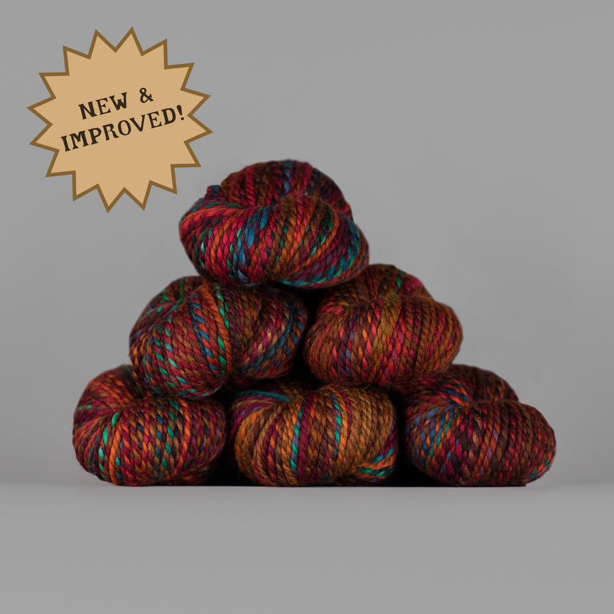 A stack of five variegated yarn skeins in rich, earthy tones with red, blue, green, and brown threads, set against a solid gray background with a gold starburst badge that reads 'NEW & IMPROVED!'.