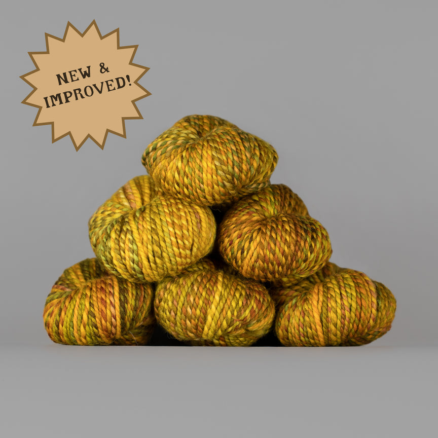 A stack of variegated yarn skeins in yellow, gold, and orange tones on a plain gray background, with a tan starburst badge in the upper left reading 'NEW & IMPROVED!'.
