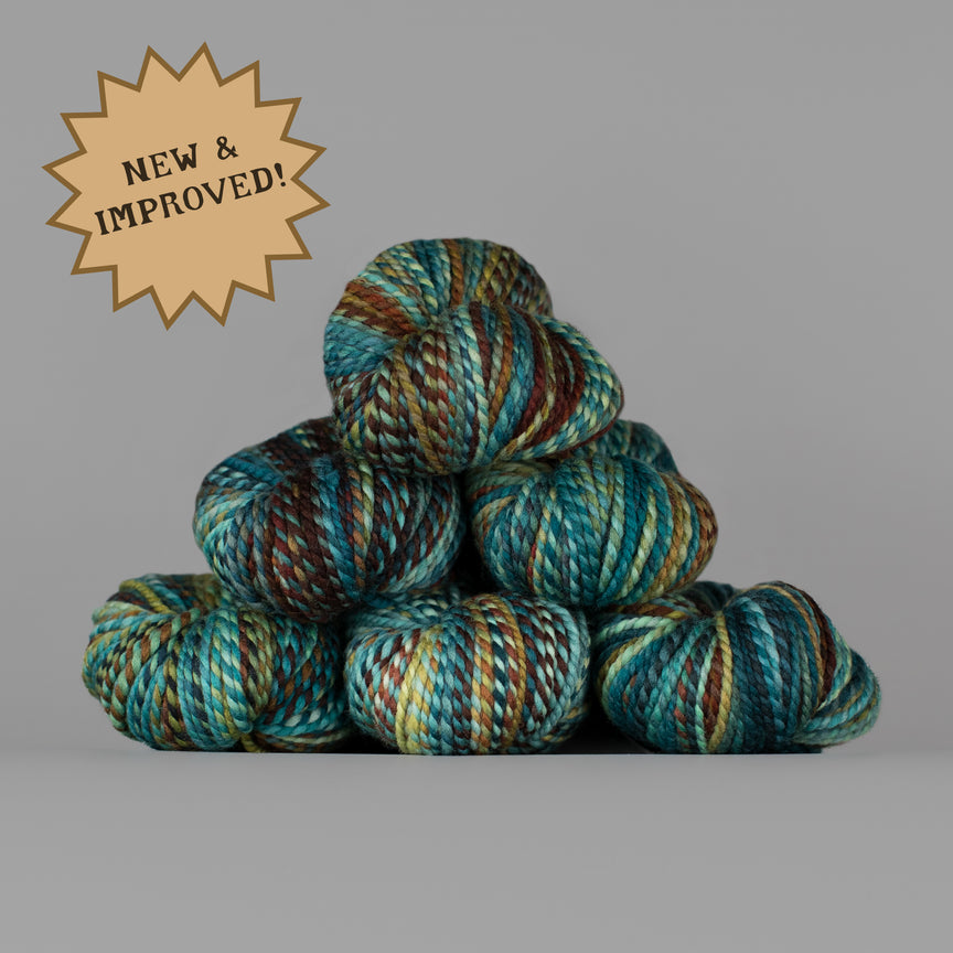 A stack of variegated yarn skeins in teal, blue, brown, and cream tones on a plain gray background, with a tan starburst badge in the upper left reading 'NEW & IMPROVED!'.